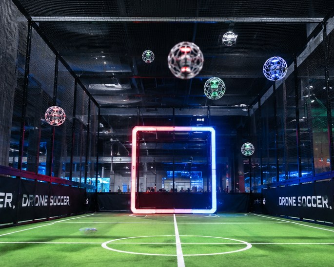 Indoor drone soccer arena with caged ball drones flying towards a lit goal ring